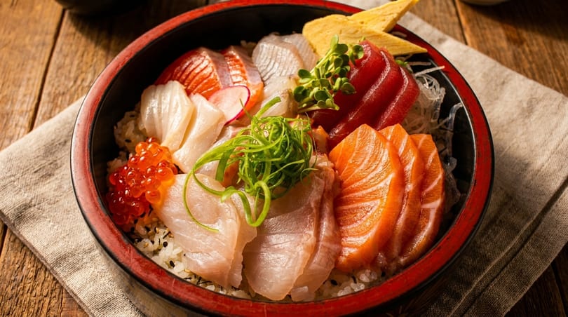 A colorful chirashi bowl with various raw fish, roe, and garnishes on rice, on a wooden table.