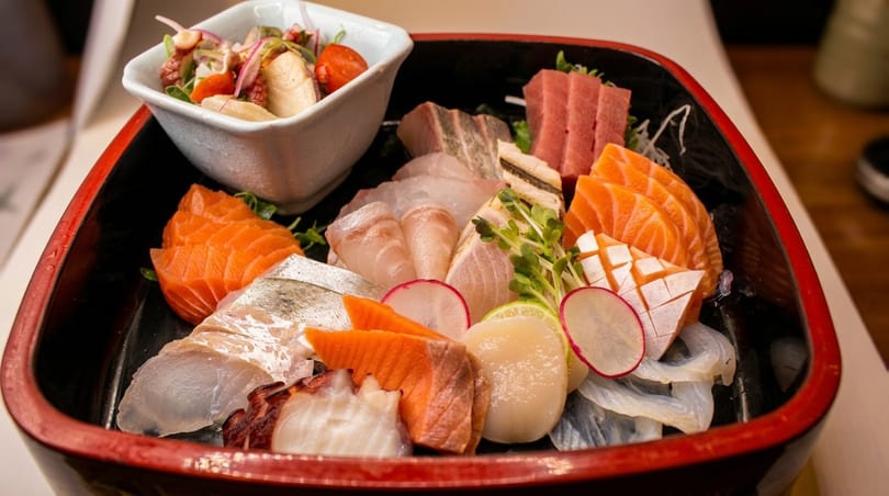 A black and red tray filled with various sashimi, including salmon, tuna, and scallops, with a side salad.