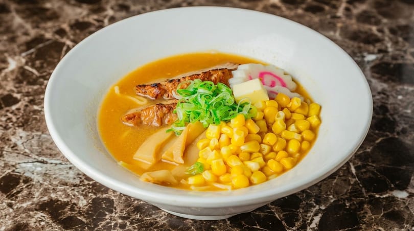 Ramen bowl with corn, green onions, fish cake, butter, and pork belly in a rich broth.